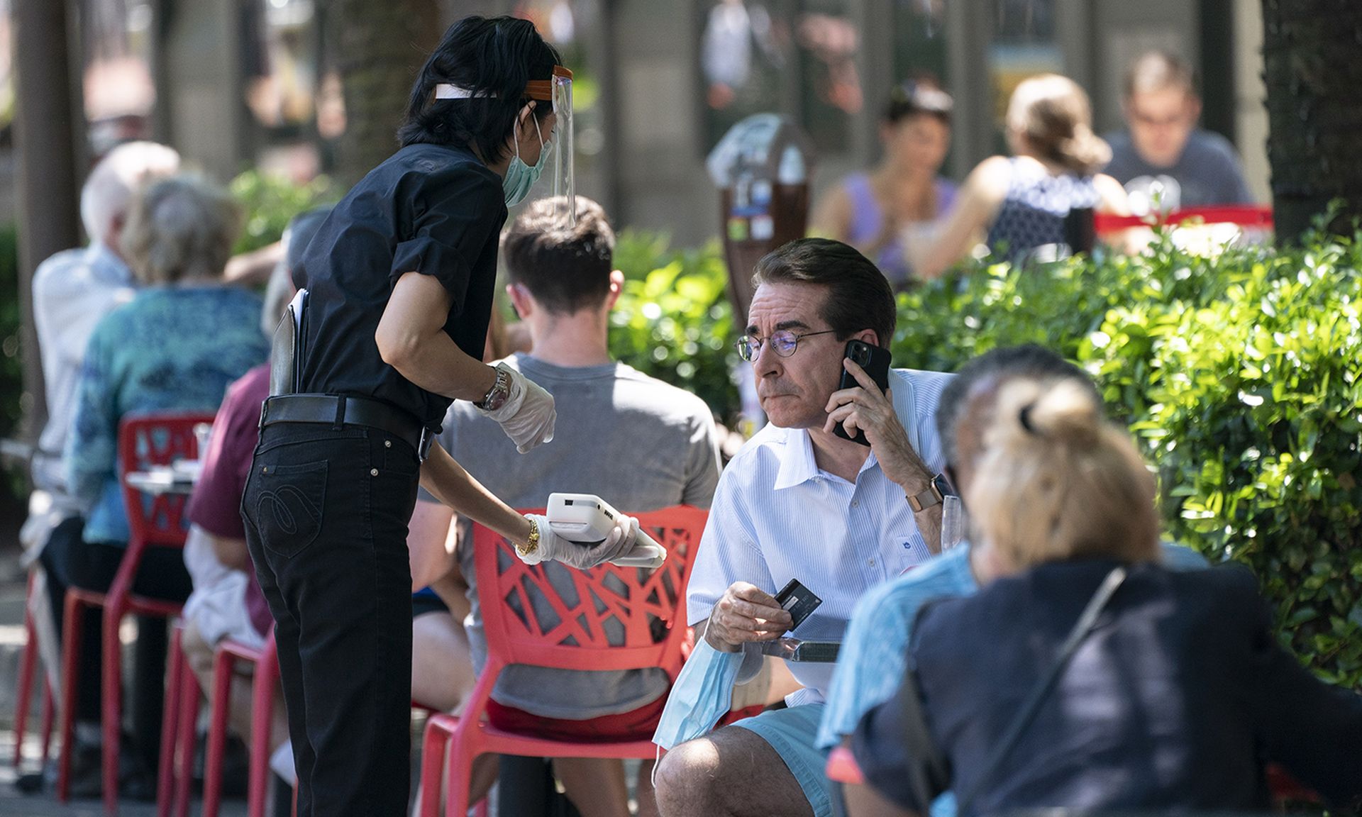 A waiter in wearing a protective face mask presents an outside diner a contactless payment system.