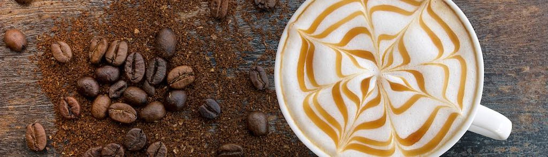 Hot Cappuccino coffee with zigzag caramel motif or spiderweb art floating on top and coffee beans with ground on wooden background. Coffee break at retro style coffee shop,t op view. (Hot Cappuccino coffee with zigzag caramel motif or spiderweb art fl