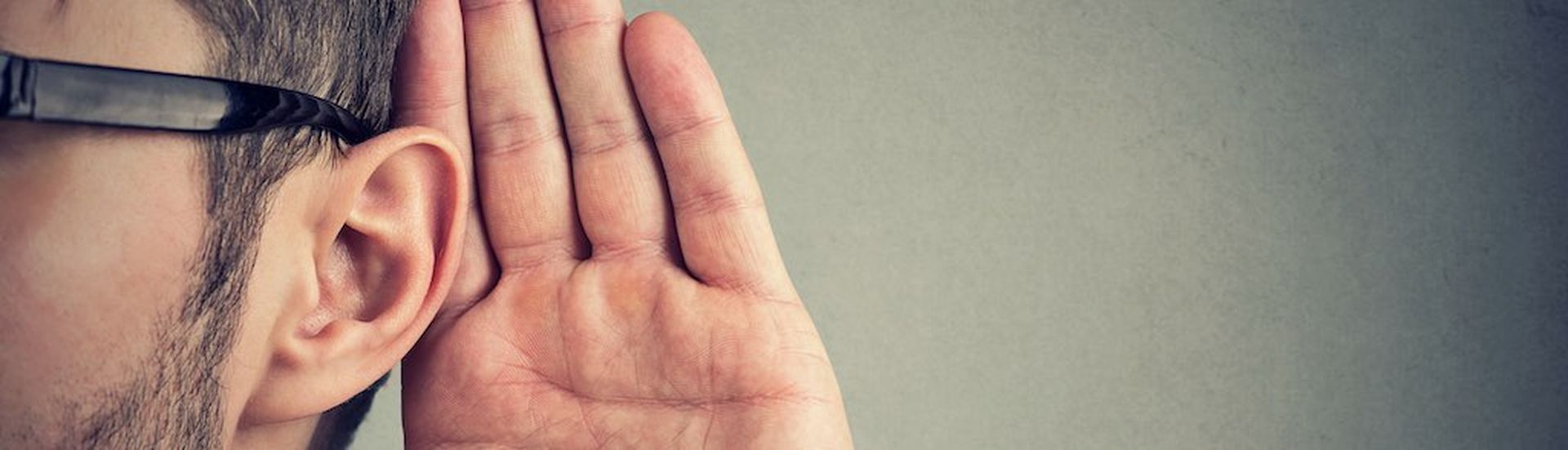 man holds his hand near ear and listens carefully isolated on gray wall background