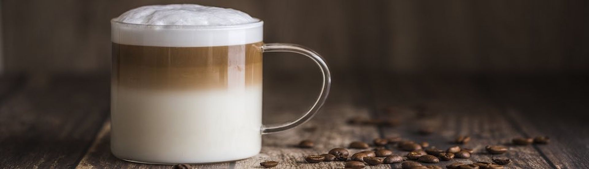 Cafe latte macchiato layered coffee in a see through glass coffee cup. The cup is on a wooden background with coffee beans on the table next to the cup. (Cafe latte macchiato layered coffee in a see through glass coffee cup. The cup is on a wooden bac