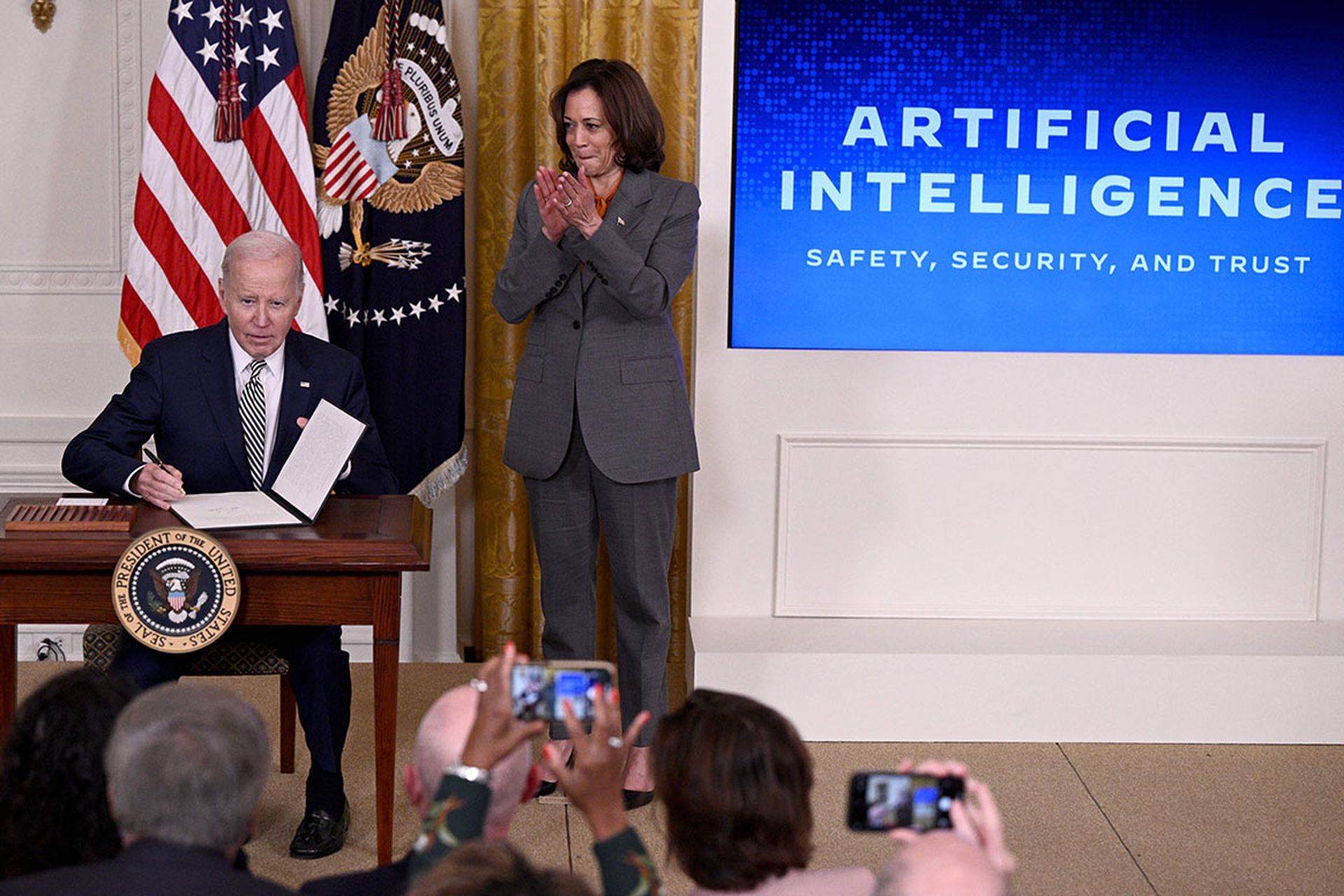 President Joe Biden sits at a table on a stage to sign an executive order while Vice President Harris applauds.