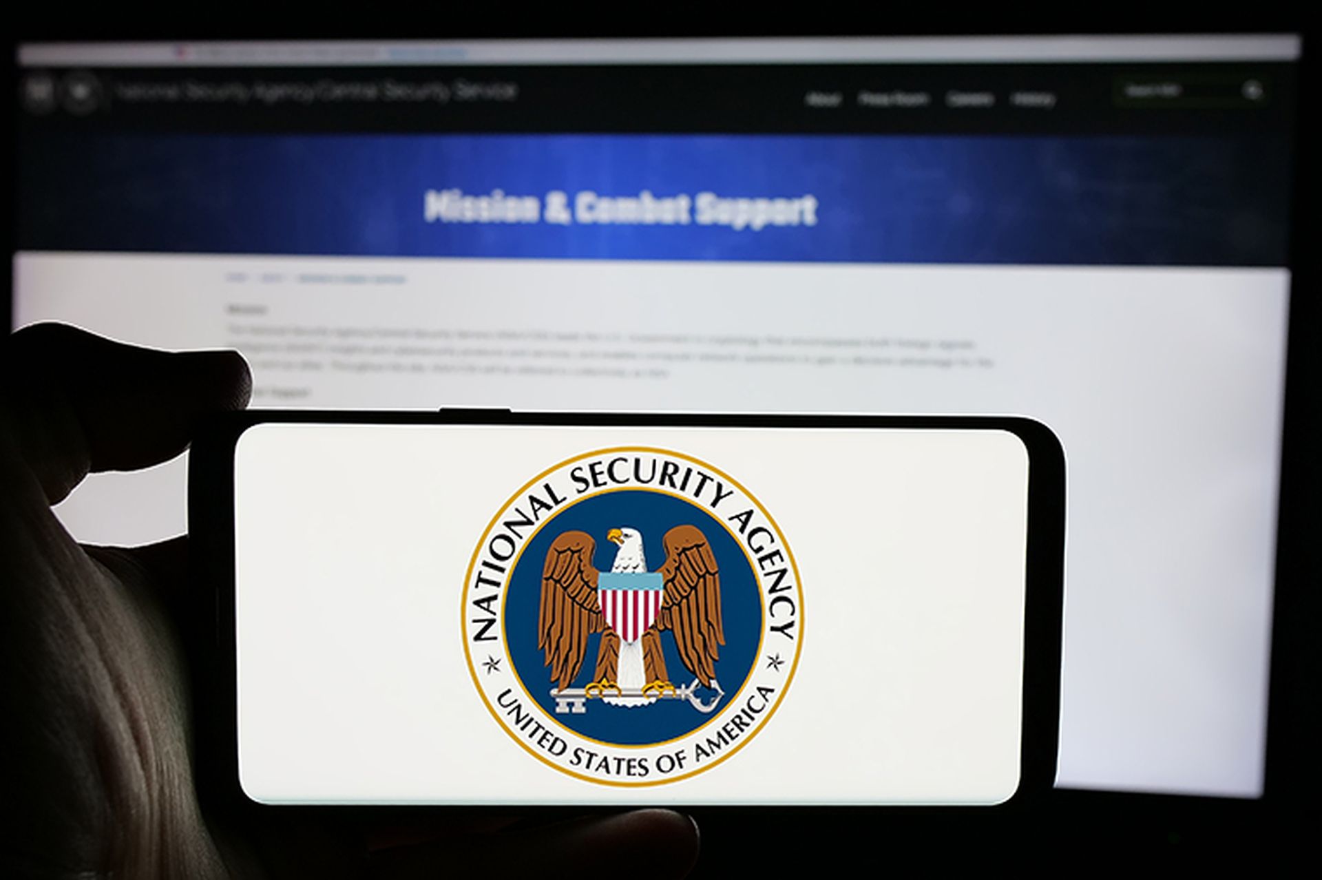 Person holding cellphone with logo of National Security Agency (NSA) on screen in front of webpage.