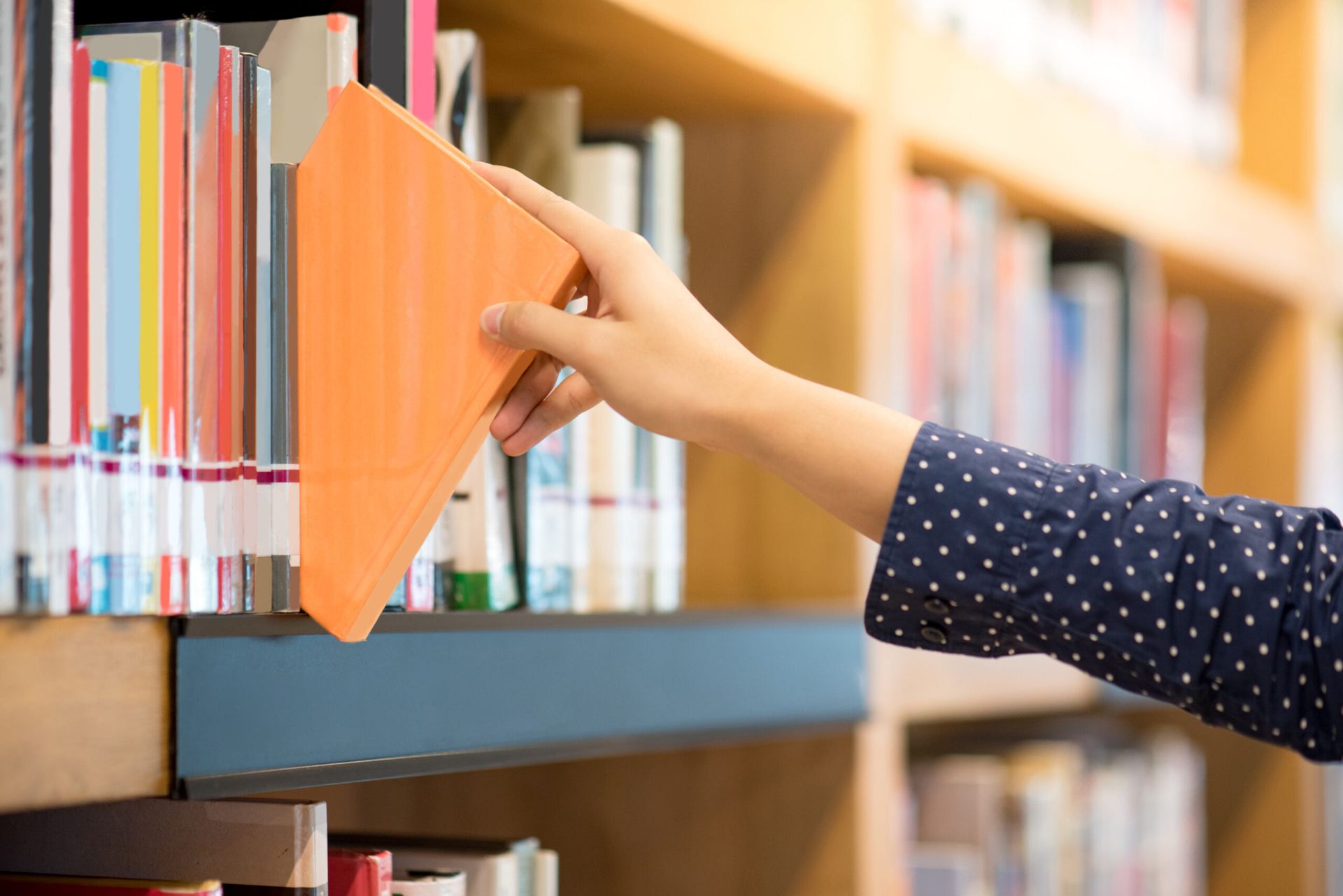 male hand choosing and picking orange book in public library, education research and self learning in university life concepts