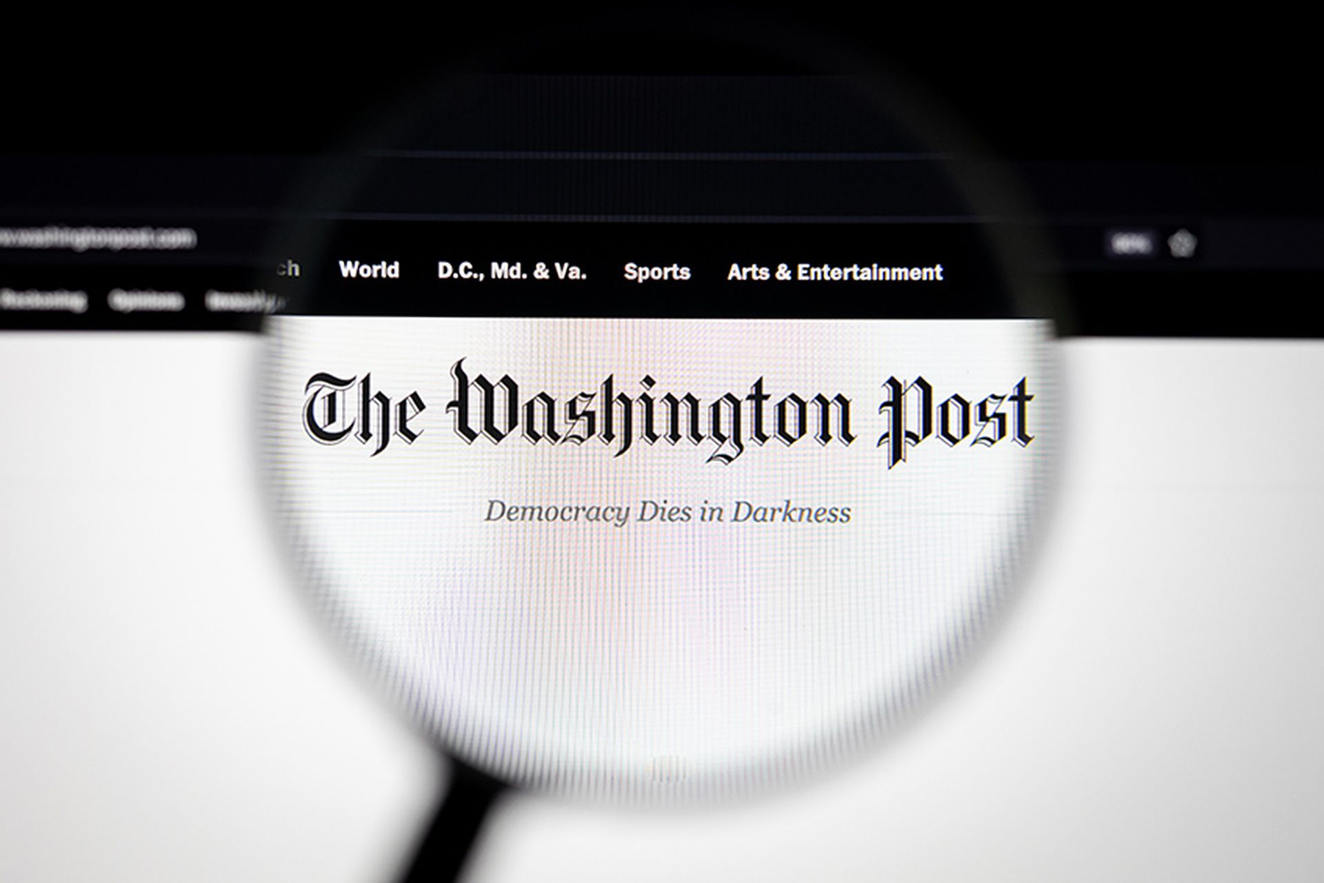 The Washington Post company logo on a website, seen on a computer screen through a magnifying glass.