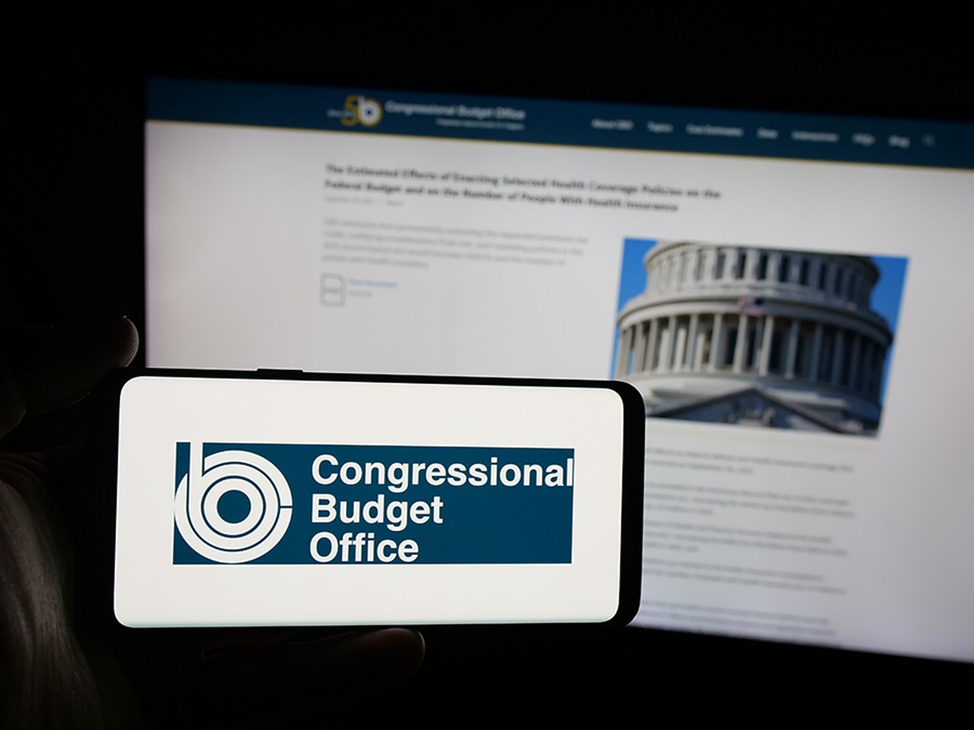 Person holding smartphone with logo of US agency Congressional Budget Office (CBO) on screen in front of website. Focus on phone display