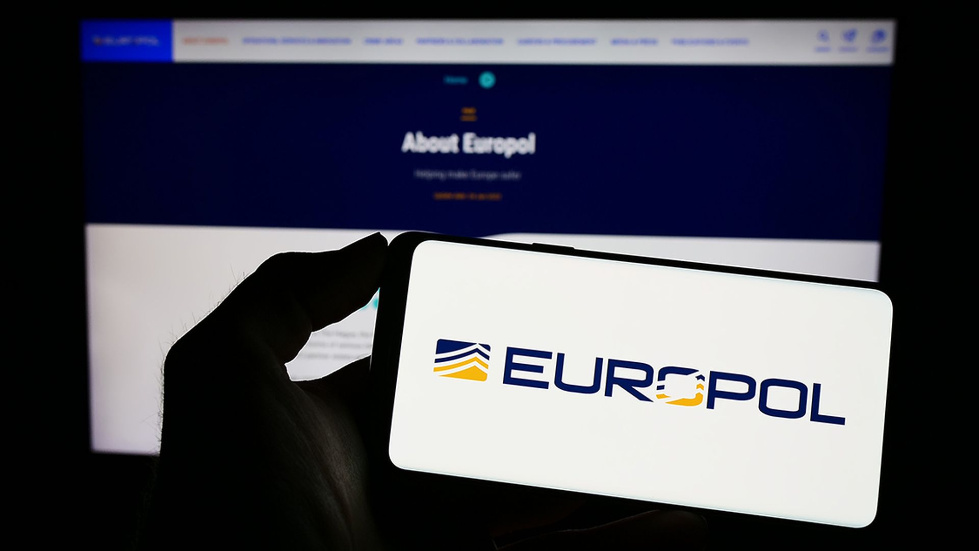Person holding cellphone with logo of EU law enforcement agency Europol on screen in front of webpage. Focus on phone display.