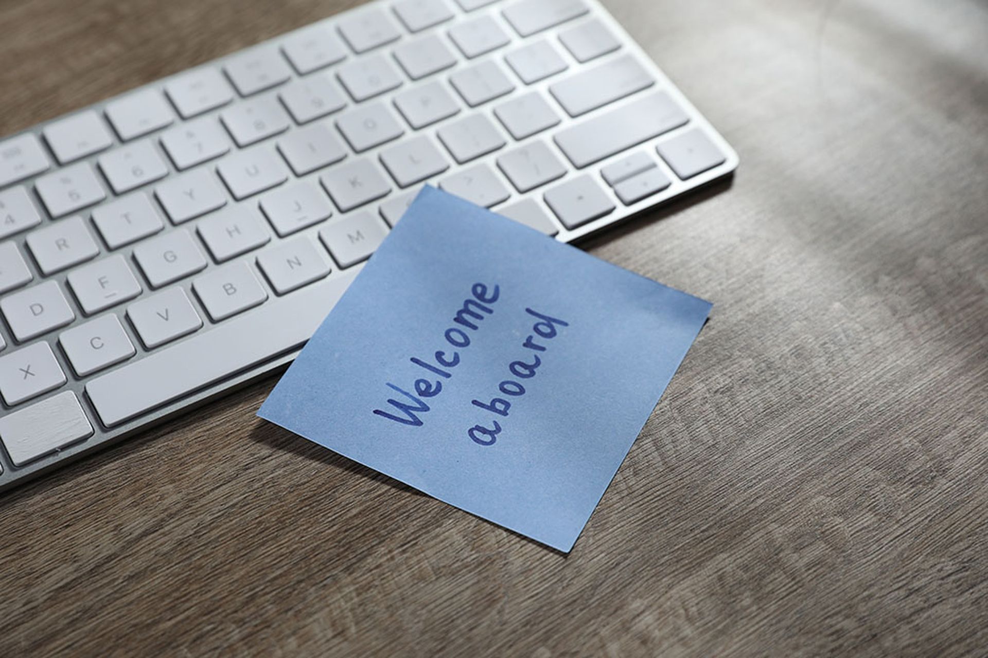 Paper note with words Welcome Aboard and computer keyboard on wooden table, closeup