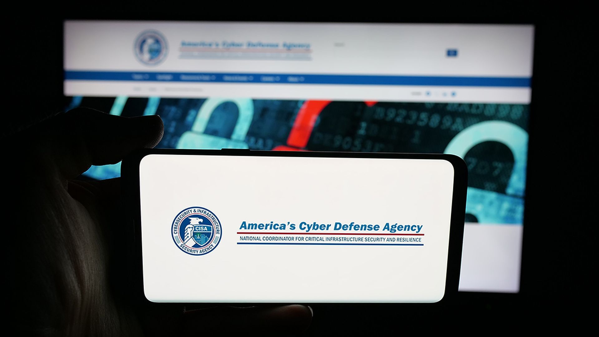 Person holding cellphone with logo of cyber security and infrastructure agency CISA on screen in front of webpage.