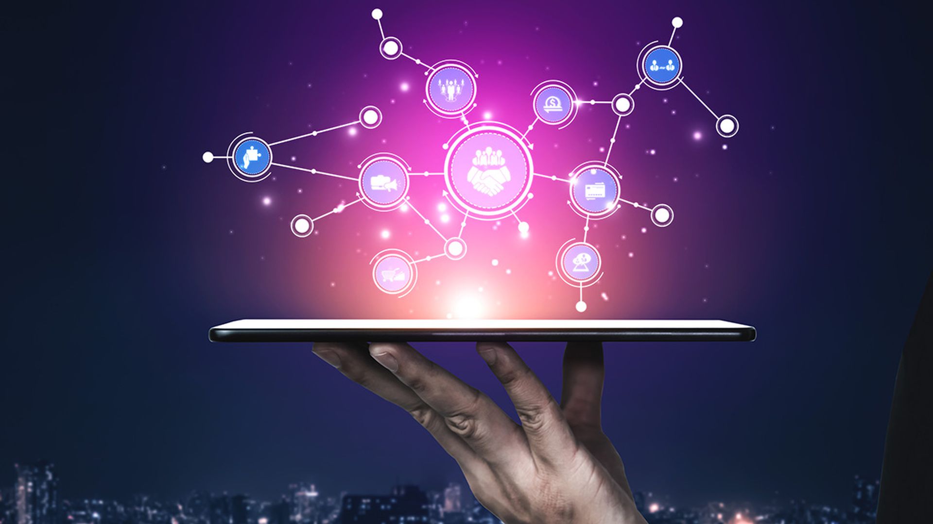 A hand holds a tablet displaying digital icons and connections against a night cityscape. This image represents technology, innovation, and modern communication concepts.