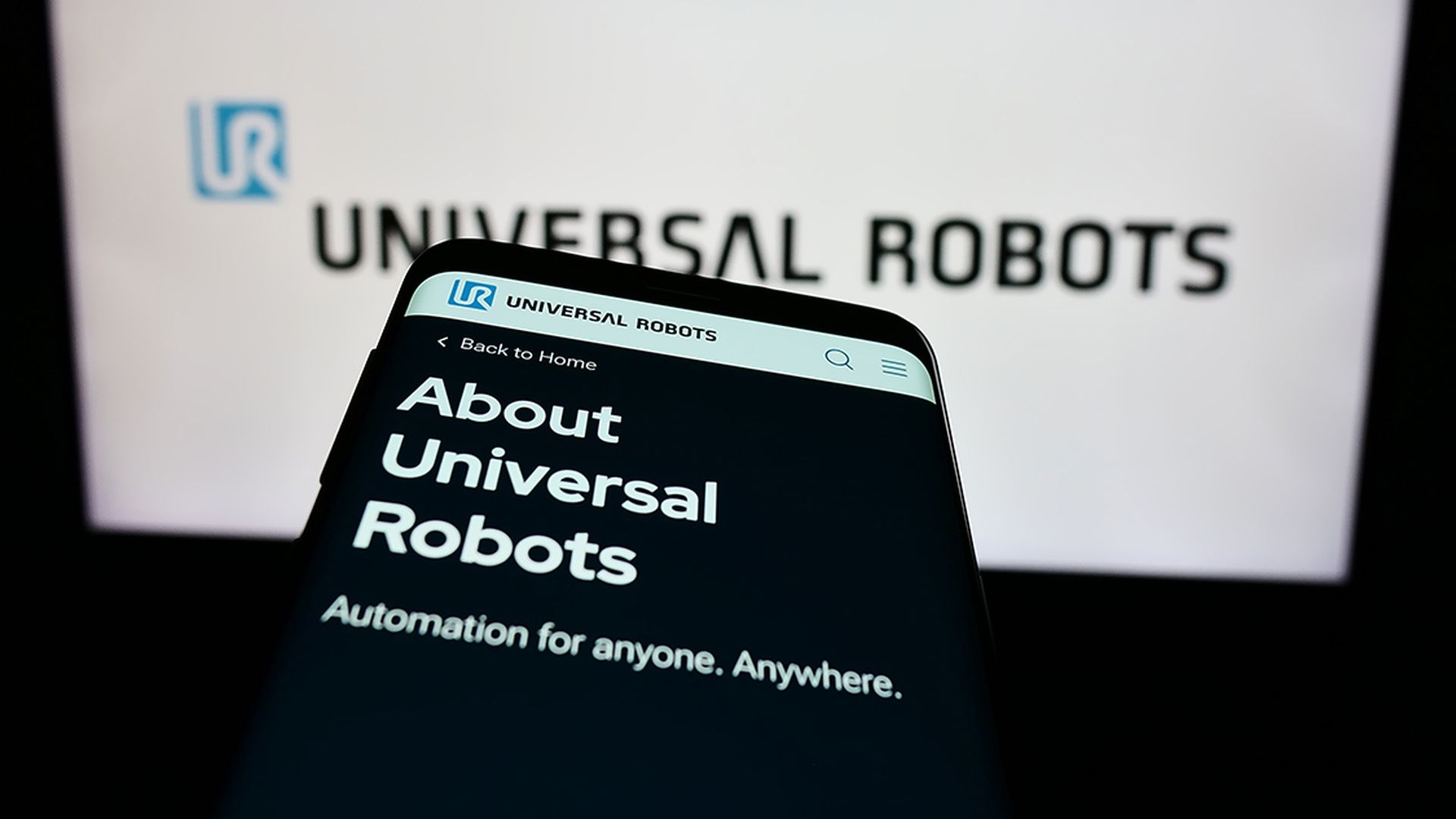 Cellphone with web page of Danish cobot company Universal Robots AS in front of business logo. Focus on top-left of phone display.