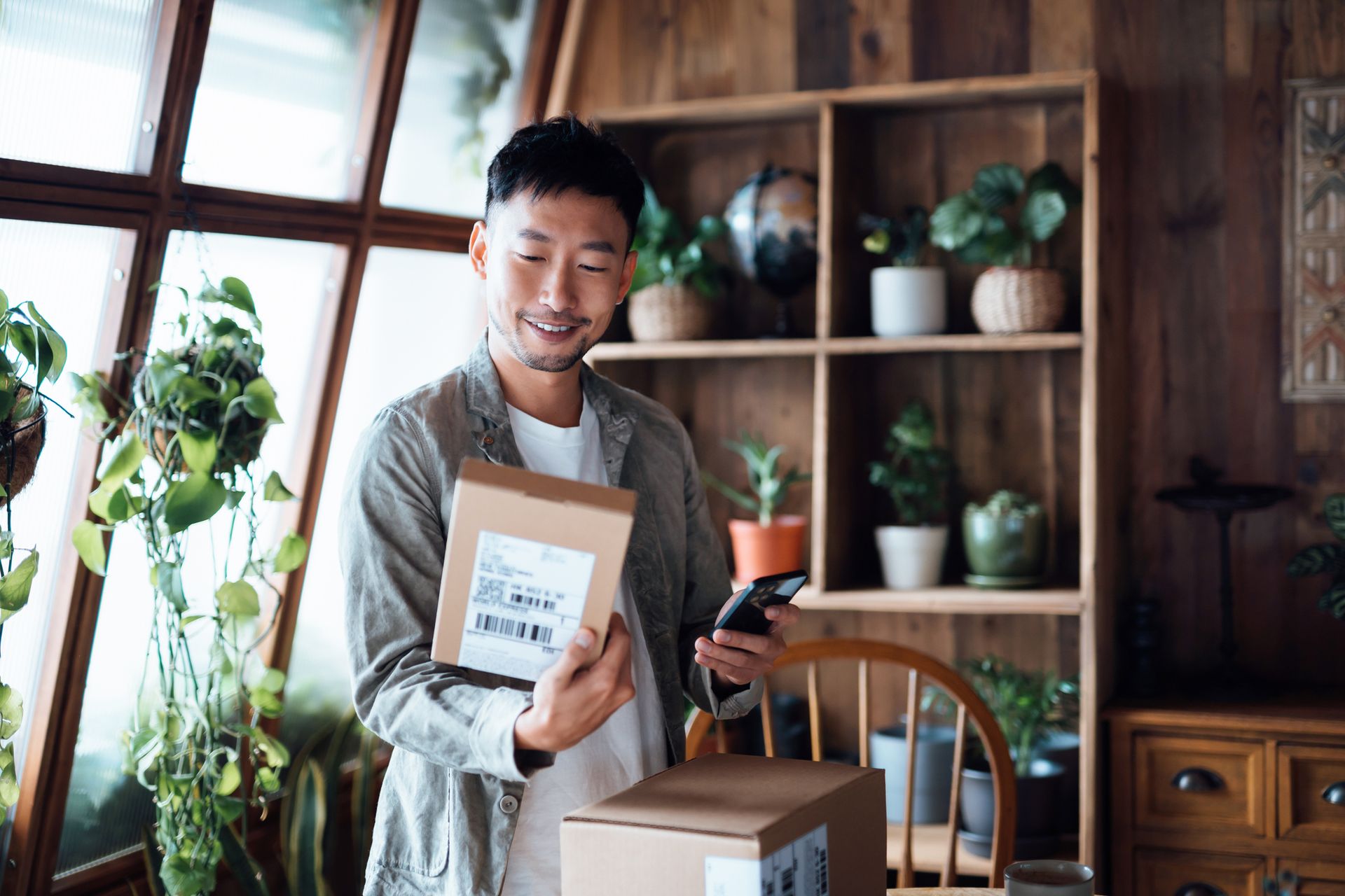 Smiling young Asian man with smartphone, receiving delivered packages from online purchases at home, can&#8217;t wait to unbox the purchases. Online shopping, enjoyable customer shopping experience