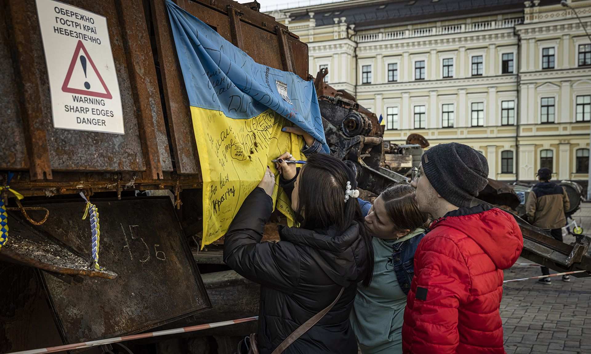 A girl writes one a Ukrainian flag as people look at an exhibition of tanks and military equipment on display.