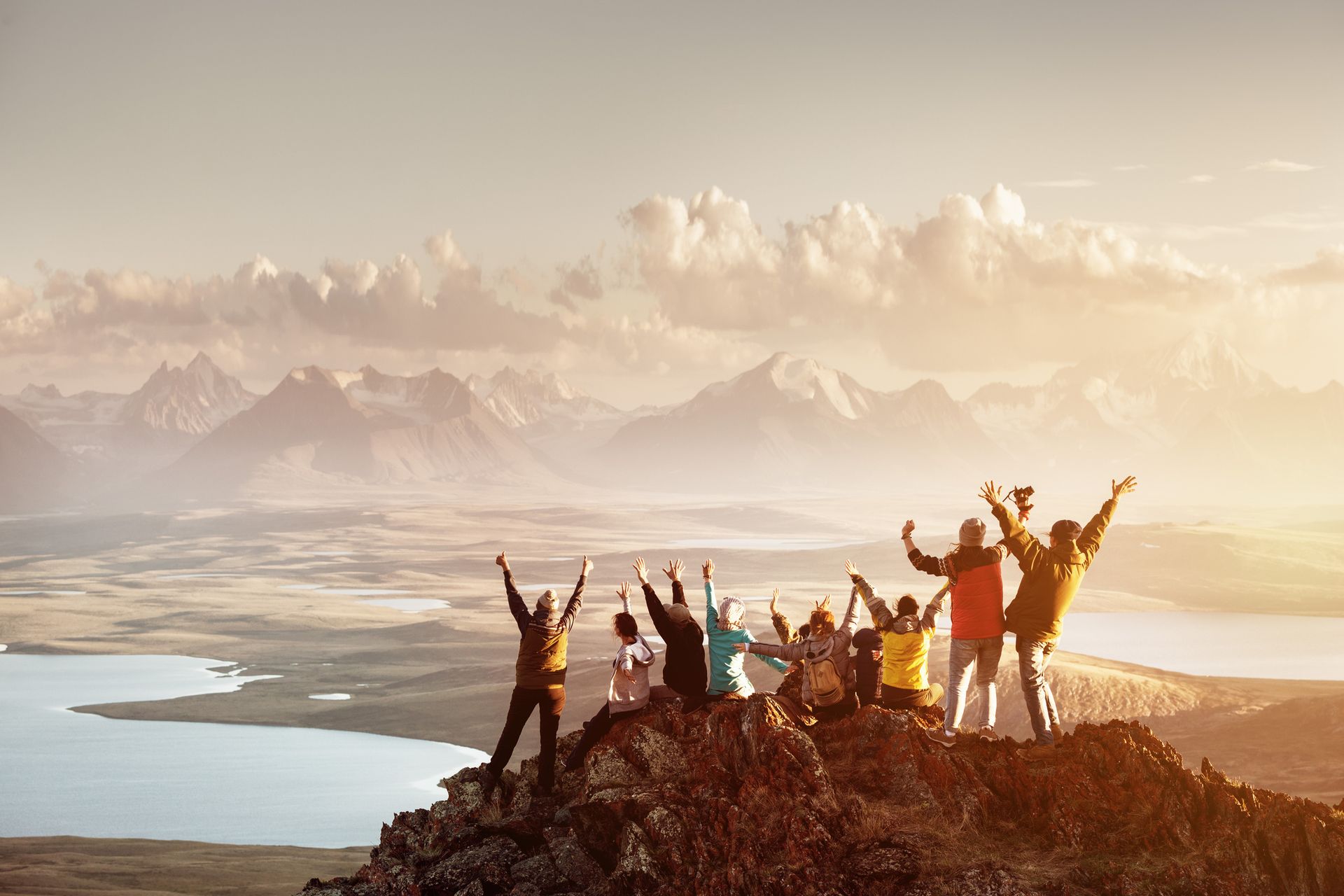 Big group of people having fun in success pose with raised arms on mountain top against sunset lakes and mountains. Travel, adventure or expedition concept