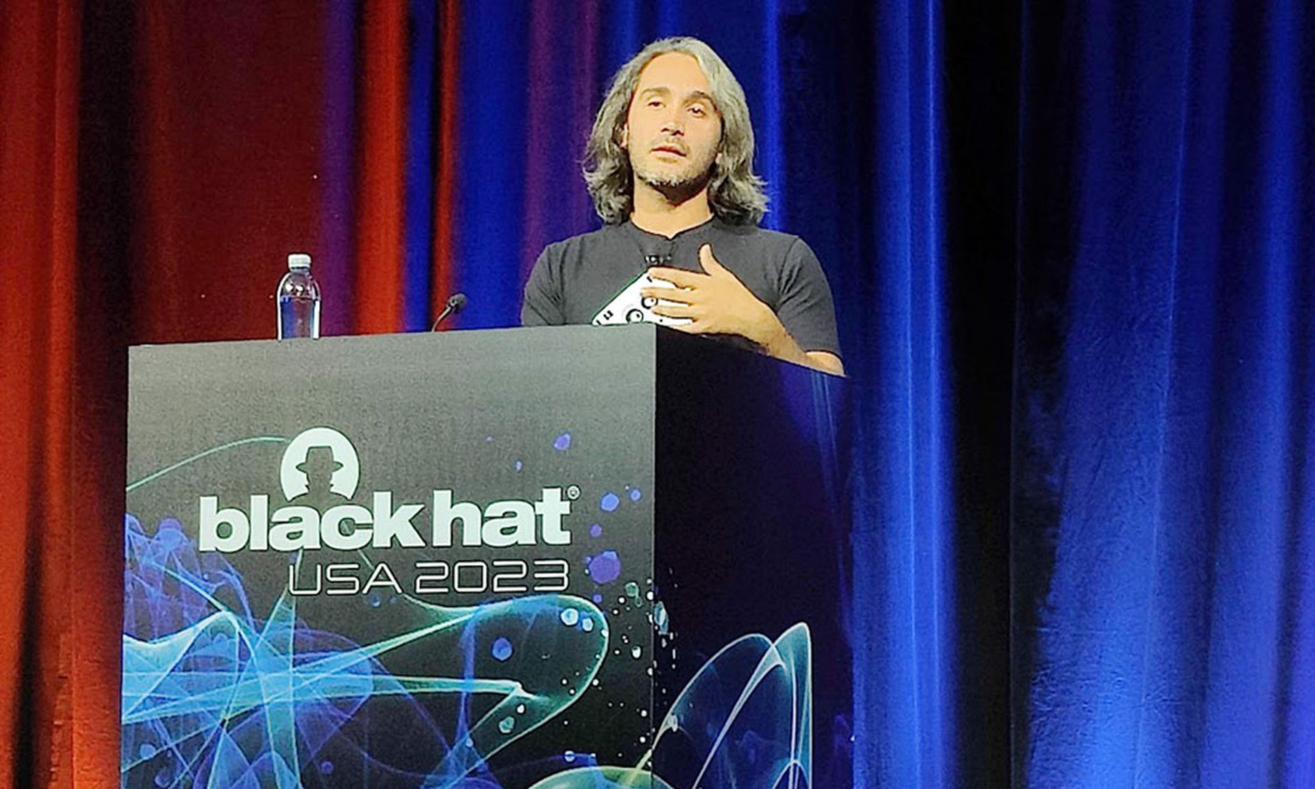 A young man with long gray hair and wearing a black T-shirt speaks at a podium labeled 'Black Hat.'