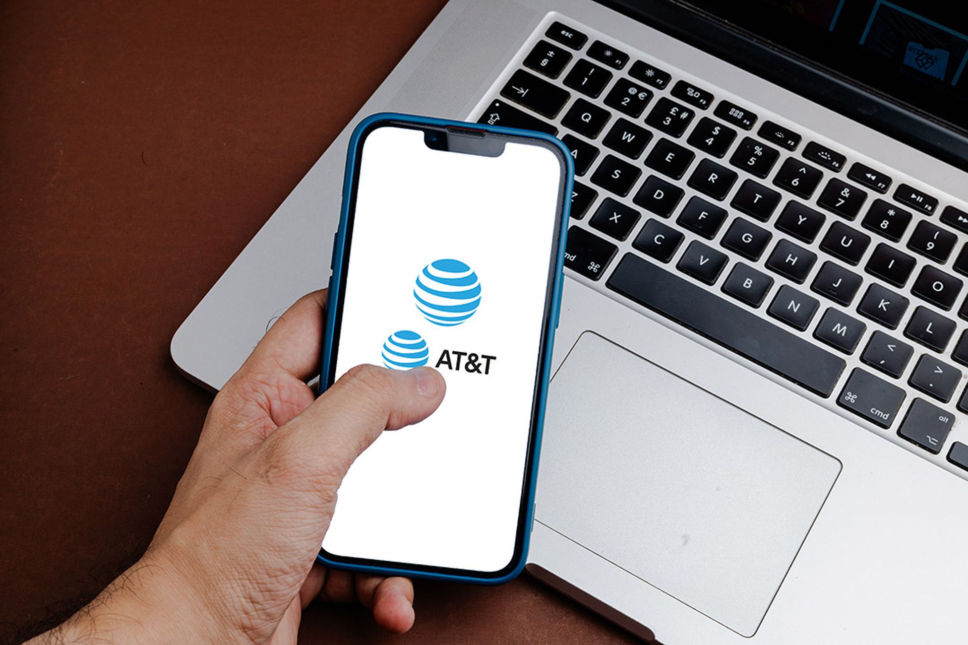 A man holds a mobile phone with the AT&T logo on the screen with a laptop in the background