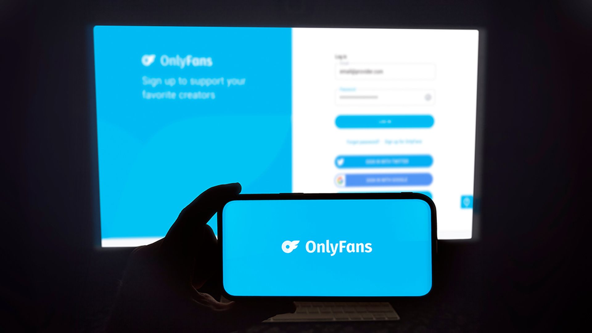 OnlyFans logo shown on a modern mobile phone. The smartphone is being held in landscape mode, in the darkness. A PC monitor with the OnlyFans website in the background.