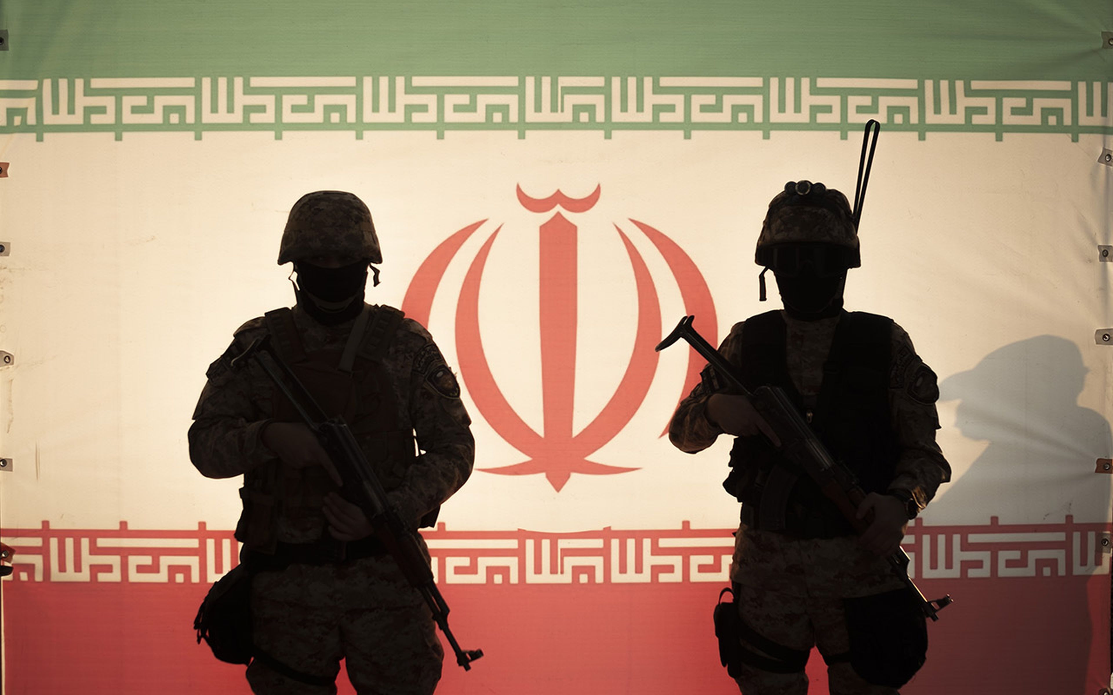 Armed military personnel stand in front of the Iranian flag