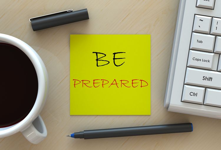 The message "Be Prepared" is written on a Post-It note on a table, in between a coffee cup and computer keyboard